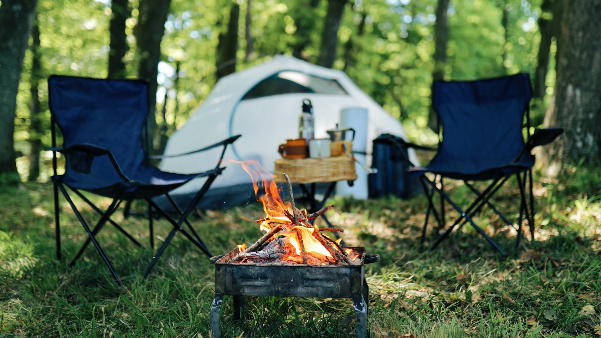 Two chairs by a small campfire with a tent and woods in the background.