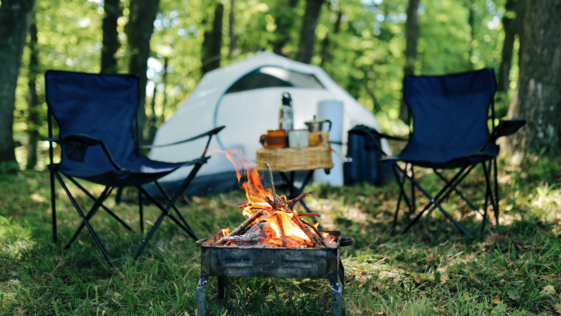 Two chairs by a small campfire with a tent and woods in the background. 